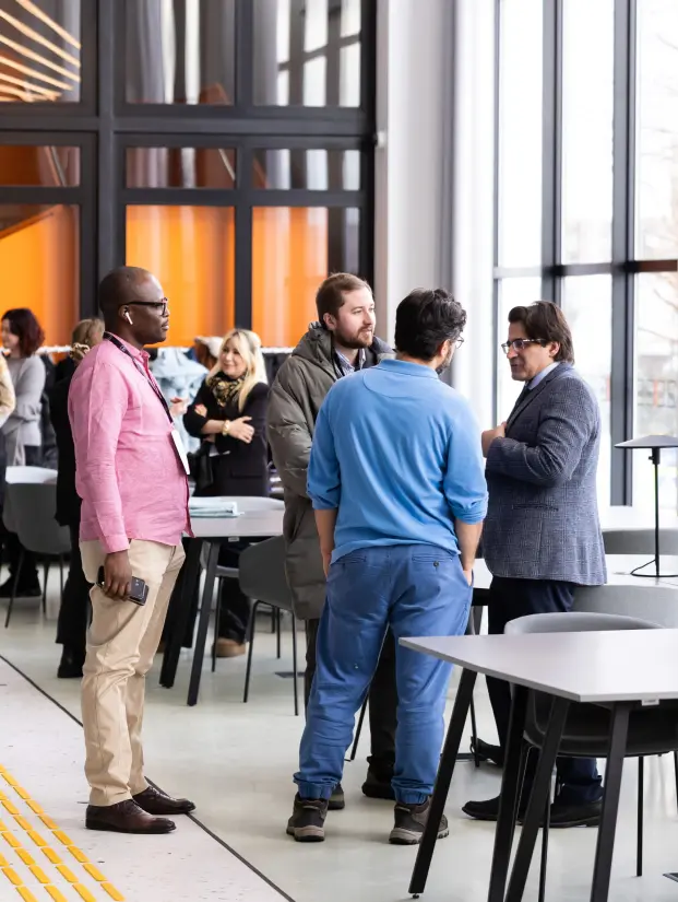 Four people in conversation in a bright modern space beside large windows, with other event participants interacting in the background. The professional setting reflects the exchange of knowledge and professional connections relevant to MBA studies.