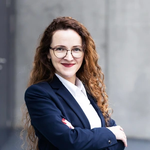 A professional-looking woman with curly brown hair and glasses, wearing a navy blue suit and a white shirt. She is smiling and appears confident. The background is a modern gray wall.