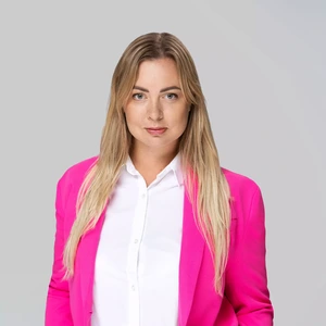 Portrait photo of a young woman with long blonde hair, wearing a bright pink blazer and a white shirt. She is looking directly at the camera with a confident, composed expression. The background is neutral, light grey.