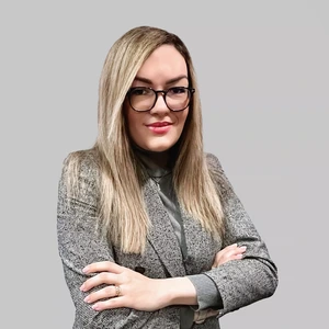 Portrait photo of a woman with long blonde hair, wearing a checkered blazer and glasses, arms crossed, looking at the camera against a light background.