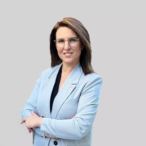 A woman with straight dark hair, wearing a light blue blazer and glasses, stands with arms crossed and smiles while looking at the camera against a light background.