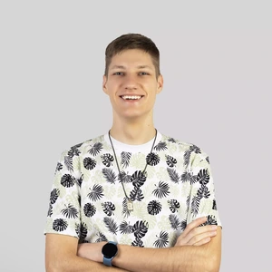 Portrait photo of a young man with short light hair, wearing a white T-shirt with a leaf pattern. He is smiling, arms crossed, and looking directly at the camera. The background is neutral, light grey.