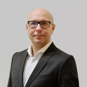 Portrait photo of a bald man wearing black glasses and an earring, dressed in a white shirt and a dark blazer. He is looking at the camera against a neutral grey background.