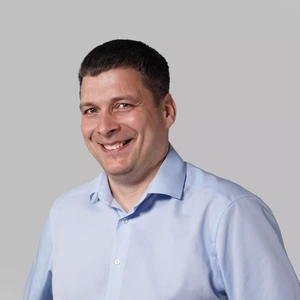 Portrait photo of a man with short dark hair, wearing a light blue shirt, smiling broadly at the camera against a light background.