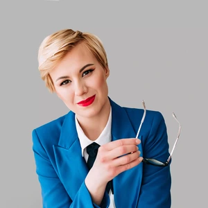 Portrait photo of a young woman with short blonde hair, dressed in a bright blue blazer, white shirt, and a black ribbon tie. She is smiling, holding glasses in her hand, and looking directly at the camera. The background is neutral grey.