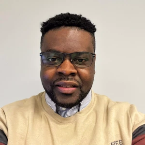 Portrait photo of a man with short curly hair and a beard, wearing glasses. He is dressed in a light-colored sweater and a striped shirt. The background is neutral light grey.