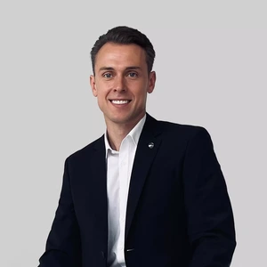 A young professional man is wearing a dark suit and white shirt, smiling at the camera against a light background.