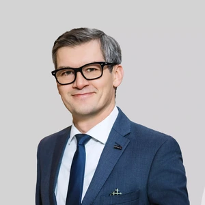 A middle-aged man with glasses and graying hair is wearing a blue suit, white shirt, and dark tie. He is smiling and looking at the camera against a light background. A pin with the text “IPMA” is attached to his blazer lapel.