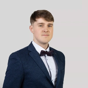 Portrait photo of a young man with short light brown hair, wearing a dark blue suit, white polka dot shirt, and a burgundy bow tie. He is looking at the camera against a neutral grey background.