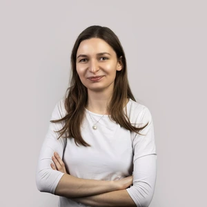Portrait photo of a woman with long brown hair, wearing a light top and a sun-shaped pendant necklace, smiling at the camera with arms crossed against a light background.