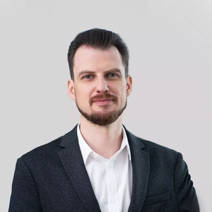Portrait photo of a man with dark slicked-back hair and a short beard, wearing a white shirt and a dark blazer. He is looking at the camera with a gentle smile, against a neutral greyish background.