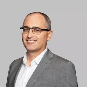 Portrait photo of a man with short grey hair, wearing glasses, a light grey suit, and a white shirt. He is looking at the camera with a broad smile, against a neutral grey background.
