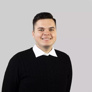 Portrait photo of a young man with short dark hair, wearing a white shirt and a black cable-knit sweater. He is smiling and looking directly at the camera, against a neutral light grey background.