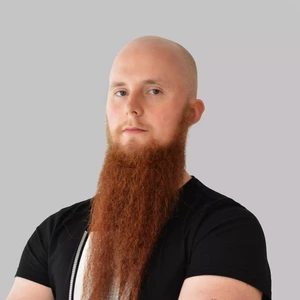 Portrait photo of a man with a shaved head and a very long, bright reddish-brown beard, wearing a black t-shirt, looking at the camera with a serious expression against a light background.