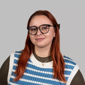 Portrait photo of a young woman with long red hair, wearing large black glasses, a blue and white striped sweater vest, and a green top. She is looking at the camera with a slight smile, against a neutral grey background.