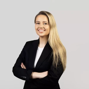 A young woman with long blonde hair, wearing a black blazer and a white top, stands with arms crossed and smiles while looking at the camera against a light background.