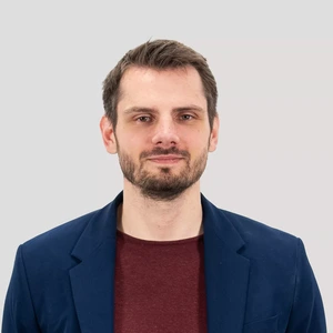 Portrait photo of a man with short brown hair and a beard, wearing a dark blue blazer and a burgundy T-shirt. He is looking at the camera against a neutral grey background.