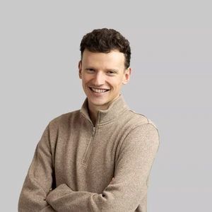 A young man with short curly hair, wearing a light brown zip-up sweater, stands with arms crossed and smiles while looking at the camera against a light background.