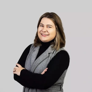 Portrait photo of a young woman with medium-length light brown hair, wearing a black turtleneck and a grey vest. She is smiling and looking at the camera, against a neutral light grey background.
