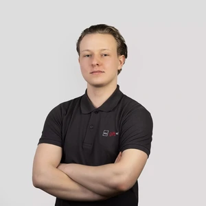 A young man with light hair, wearing a dark polo shirt with the “ktu gifted” logo, stands with arms crossed and looks at the camera with a neutral expression against a light background.