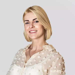 Portrait photo of a blonde woman with short hair, wearing a light sheer blouse with decorative dots, smiling at the camera against a light background.