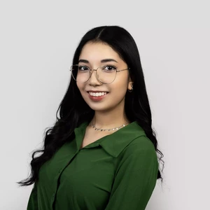 Portrait photo of a young woman with long wavy dark hair, wearing a green blouse, glasses, and a silver necklace. She is looking at the camera and smiling, with a light neutral background.
