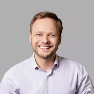 A blond man smiling, wearing a light-colored shirt, photographed against a light background.