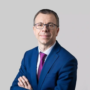 Portrait photo of an older man wearing glasses, a dark blue suit, a patterned light shirt, and a fuchsia tie. He is looking at the camera with a gentle smile, against a neutral light grey background.