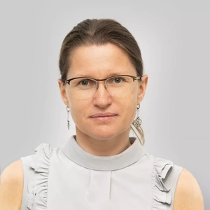 Portrait photo of a woman wearing glasses with her hair tied back. She is dressed in a sleeveless light grey blouse with pleated details. She is looking directly at the camera, with a light grey background.