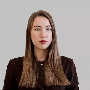 A serious young woman with long brown hair, wearing a dark blazer, posing against a light background.