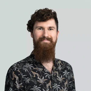 A man with curly dark hair and a long beard is wearing a black tropical-patterned shirt and looks at the camera with a gentle smile against a light background.