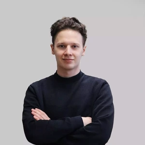A young man with short dark hair, wearing a black sweater, stands with arms crossed and looks at the camera with a slight smile against a light background.