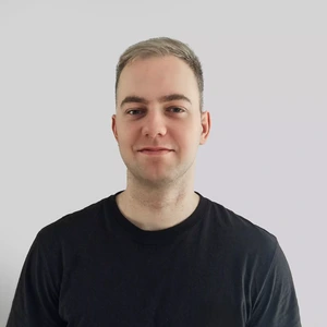 A young man with short light hair and a black t-shirt looking at the camera with a neutral expression, set against a light background.