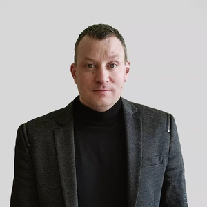 Portrait photo of a man with short dark hair, wearing a black turtleneck and a dark textured blazer. He is looking directly at the camera with a serious expression. The background is neutral, light grey.