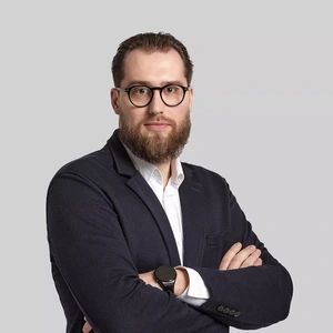 A serious man with a beard and glasses, wearing a dark blazer and white shirt, standing with arms crossed against a light background.