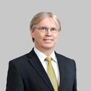 Portrait photo of a middle-aged man with light hair, wearing a black suit, white shirt, and a yellow striped tie. He is looking at the camera with a slight smile, against a light grey neutral background.