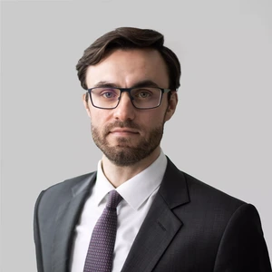 Portrait photo of a man with dark hair and a beard, wearing glasses, a dark suit, a white shirt, and a patterned purple tie. He is looking at the camera with a serious expression, against a light grey background.