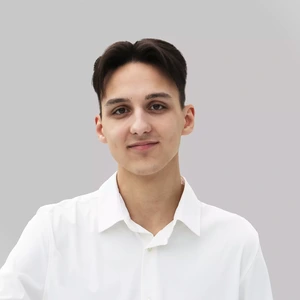 Portrait photo of a young man with dark hair, wearing a white shirt, slightly smiling and looking at the camera against a light background.