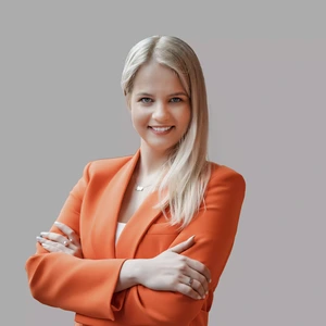 A young woman with long blonde hair, wearing a bright orange blazer, stands with arms crossed and smiles while looking at the camera against a grey background.