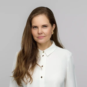 Portrait photo of a woman with long brown hair, wearing a white shirt with gold buttons and gold hoop earrings. The background is light grey, and she is looking at the camera with a subtle smile.