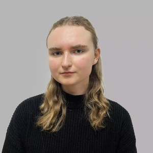 Portrait photo of a young woman with wavy blond hair, wearing a black high-neck sweater. She is looking at the camera with a serious expression, against a neutral greyish background.
