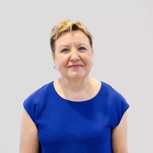 Portrait photo of an older woman with short light hair, wearing a bright blue blouse, looking at the camera with a gentle smile against a light background.