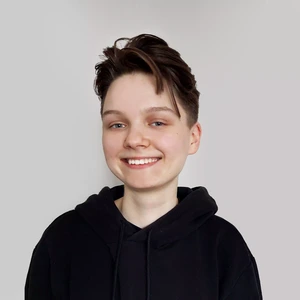 Portrait photo of a smiling young woman with short, tousled hair, wearing a black hoodie. She is looking at the camera against a light, neutral background.