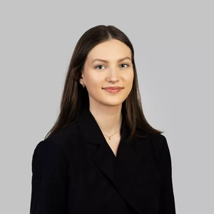 Portrait photo of a young woman with long dark brown hair, wearing a black blazer, delicate earrings, and a thin necklace. She is looking at the camera with a slight smile, against a neutral light grey background.