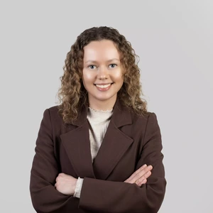 Portrait photo of a woman with curly blonde hair, wearing a brown blazer and a light turtleneck. She is smiling with arms crossed, against a light grey background.