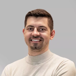 Smiling man with short hair and light facial hair, wearing a light-colored turtleneck sweater, standing against a gray background.