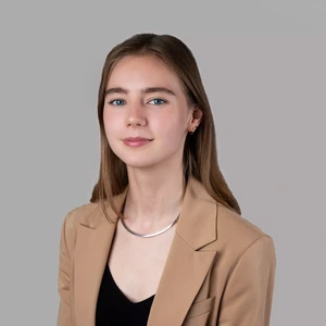 Portrait photo of a young woman with long light brown hair, wearing a beige blazer and a black top. She is looking at the camera against a neutral gray background. A silver necklace is visible around her neck.