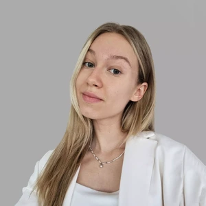 Portrait photo of a young woman with long blonde hair, wearing a white blazer and a white top. She is looking at the camera with her head slightly tilted. The background is a neutral gray. A delicate necklace with a pendant is visible around her neck.