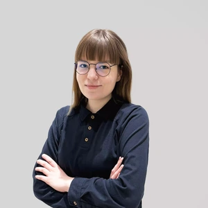 Young woman with brown hair and bangs, wearing dark blue buttoned shirt and glasses, standing with arms crossed against a gray background.