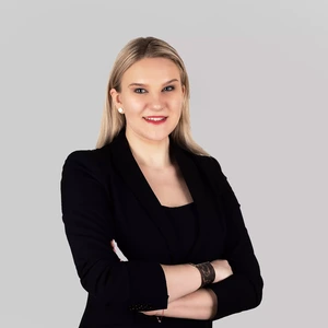 Portrait photo of a young woman with long blonde hair, wearing a black blazer and top. She is standing with arms crossed, smiling, and accessorized with a bracelet and pearl-like earrings. The background is neutral light grey.